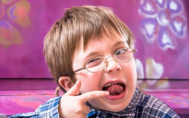 A young boy with glasses and a finger in his mouth.