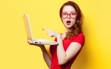 A woman wearing glasses is holding a laptop over a yellow background.