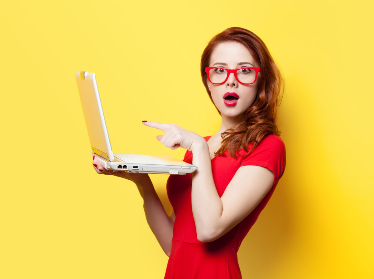 A woman wearing glasses is holding a laptop over a yellow background.