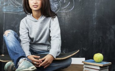 A young woman sitting on a desk with an apple and a skateboard in front of a chalkboard.