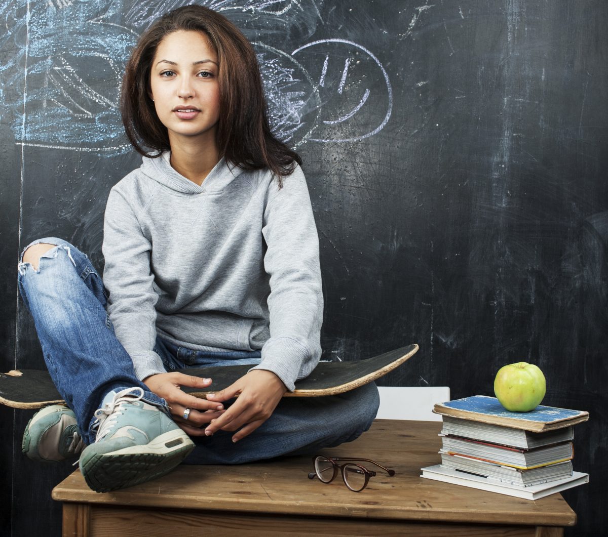A young woman sitting on a desk with an apple and a skateboard in front of a chalkboard.